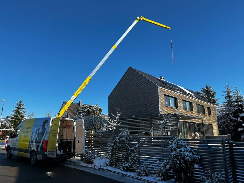 Ein Kollektormontageservice Fahrzeug mit ausgefahrenem gelben Hebearm steht vor einem modernen Wohnhaus mit Solarthermie auf dem Dach. Der Himmel ist blau, und die Umgebung ist winterlich mit schneebedeckten Bäumen und Zäunen