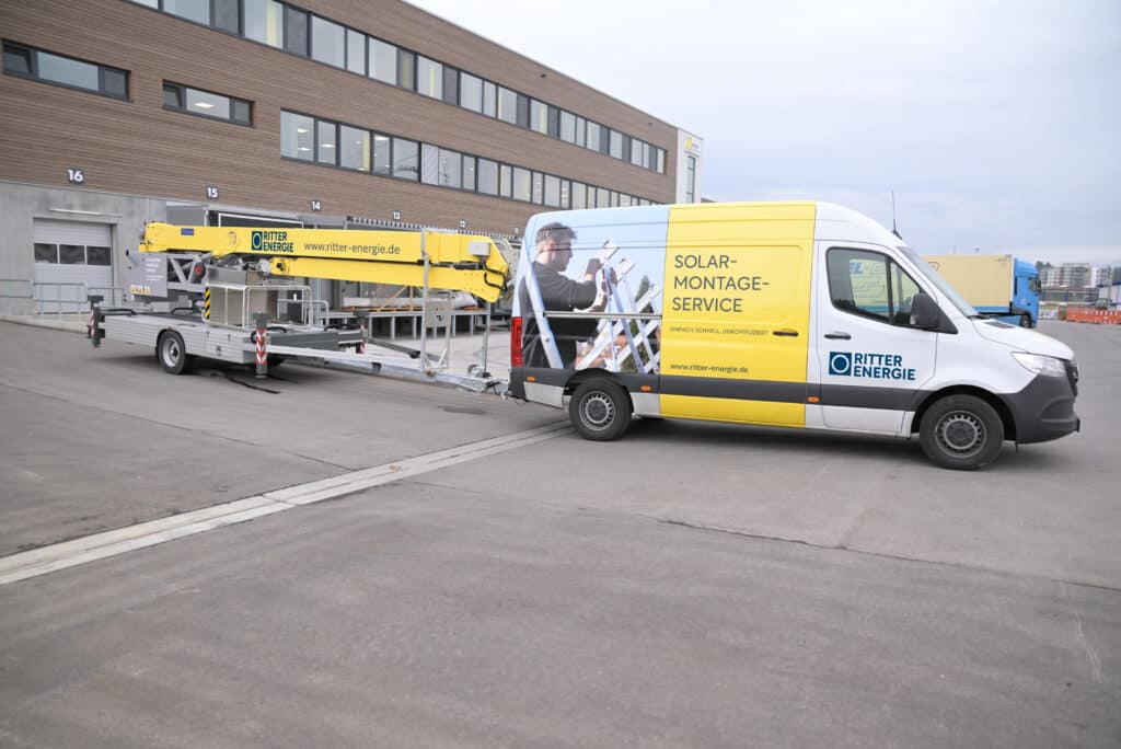 Ein Transporter von Ritter Energie mit gelb-weißer Folierung steht auf einem Industriegelände. Auf der Fahrzeugseite steht: ‚SOLAR-MONTAGE-SERVICE – Einfach. Schnell. Unkompliziert und dem Ritter-Energie-Logo. Der Transporter zieht einen Anhänger mit einer großen gelben Montageplattform