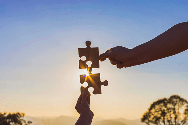 Zwei Hände halten passende Puzzlestücke vor einem Sonnenuntergang. Die Sonne scheint genau durch die Öffnung des Puzzles, während der Himmel in sanften Blau- und Orangetönen leuchtet